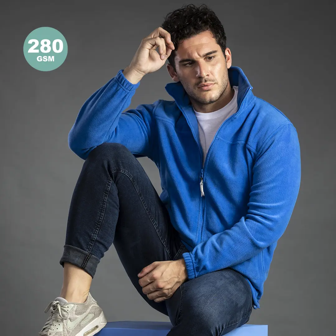 Hombre sentado con chaqueta de forro polar azul (280 GSM), vaqueros y zapatillas deportivas sobre fondo neutro