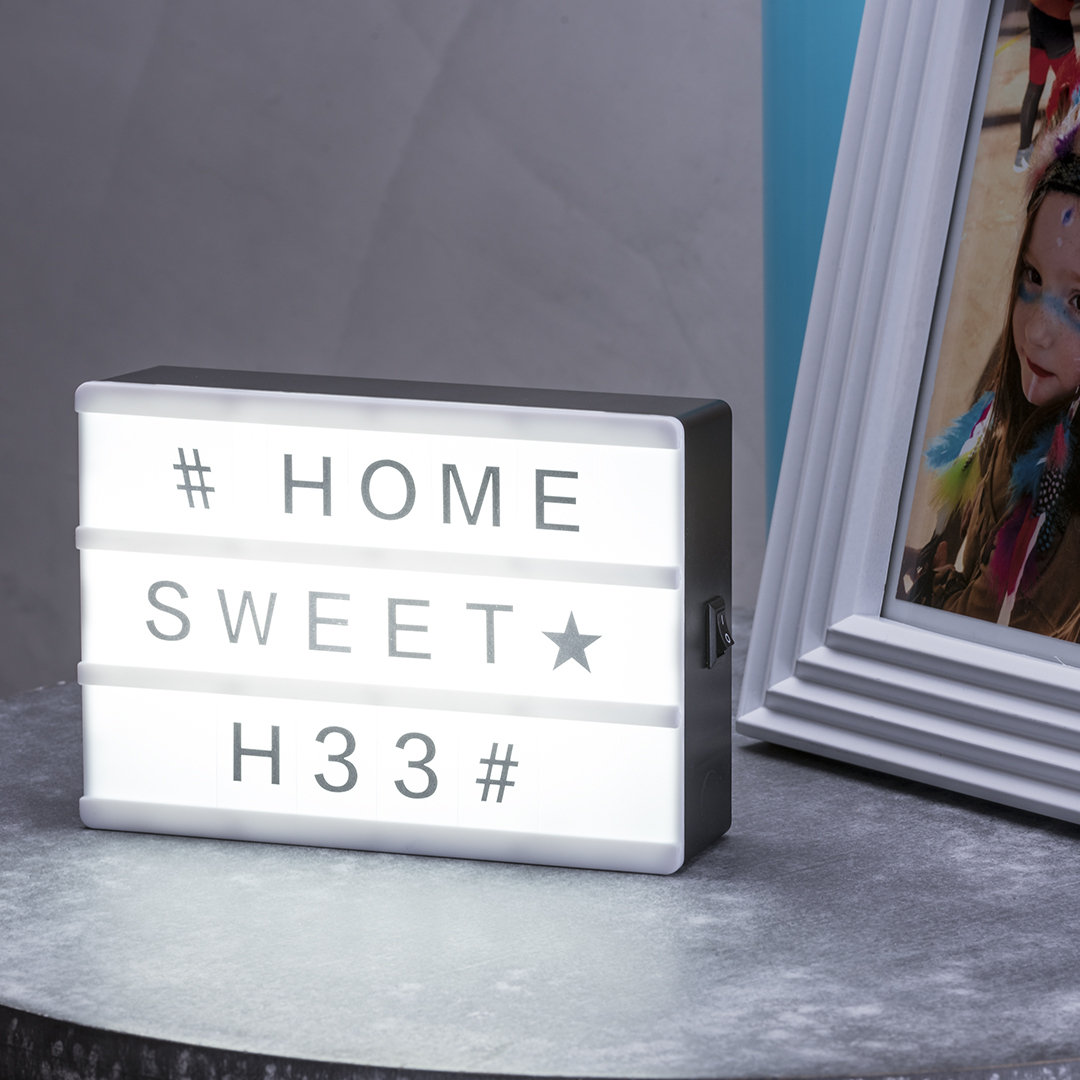 Letrero luminoso tipo lightbox con el mensaje # HOME SWEET H33# sobre una mesa junto a un marco de fotos.