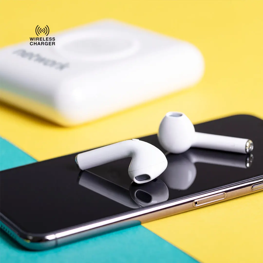 smartphone con pantalla apagada apoyado sobre fondo bicolor y auriculares inalámbricos blancos colocados encima, con cargador inalámbrico difuminado al fondo
