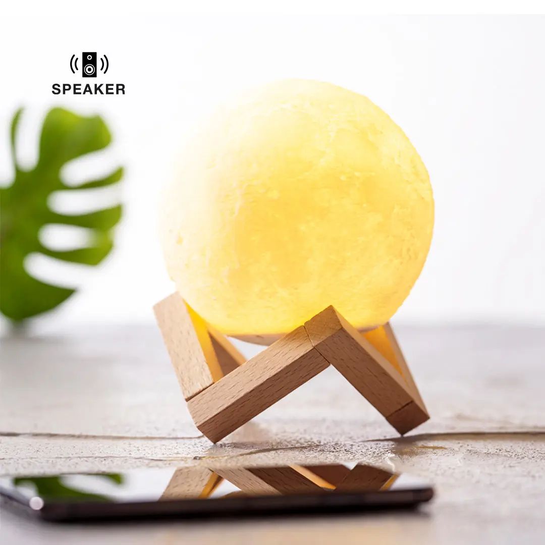 Lámpara LED en forma de luna sobre soporte de madera con altavoz Bluetooth y smartphone reflejado.