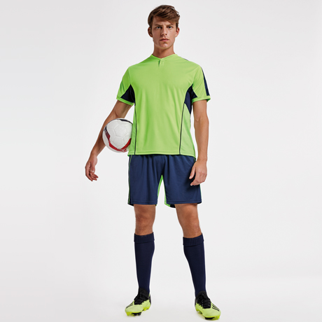 Hombre con equipación de fútbol verde y azul sosteniendo un balón sobre fondo blanco