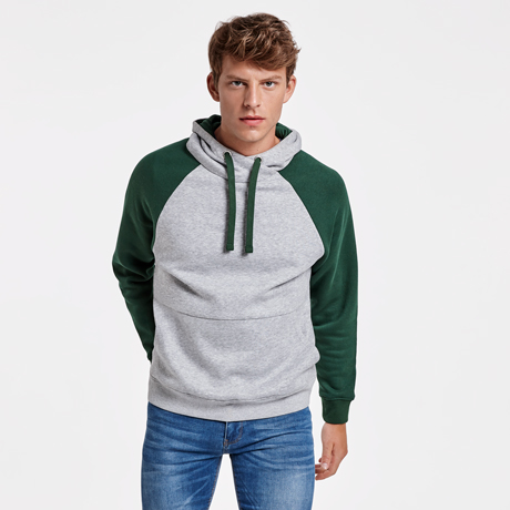 Hombre joven de pie con sudadera con capucha gris y mangas verdes sobre fondo blanco.