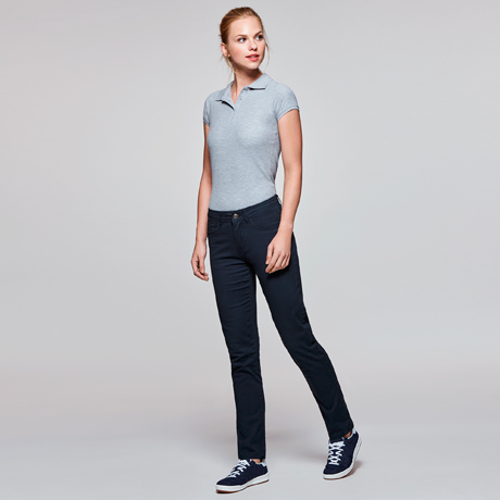 Mujer joven de pie con polo gris, pantalones oscuros y zapatillas deportivas sobre fondo neutro