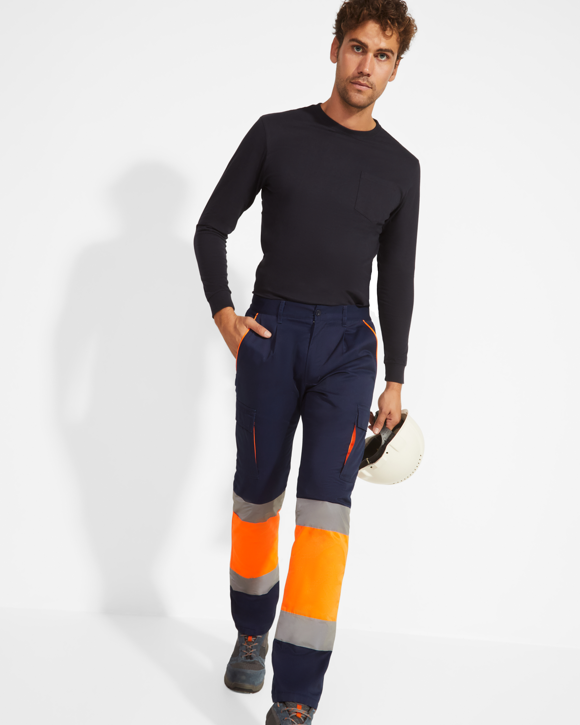 Hombre de pie con camiseta negra, pantalones de alta visibilidad naranja y azul marino con franjas reflectantes, sujetando un casco de obra blanco.