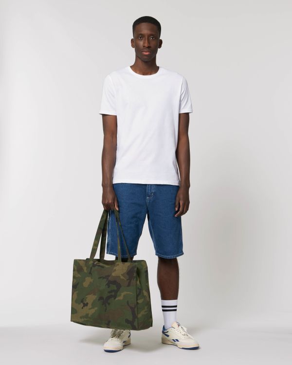 hombre joven de pie con camiseta blanca y pantalones cortos vaqueros sosteniendo un bolso tote de camuflaje