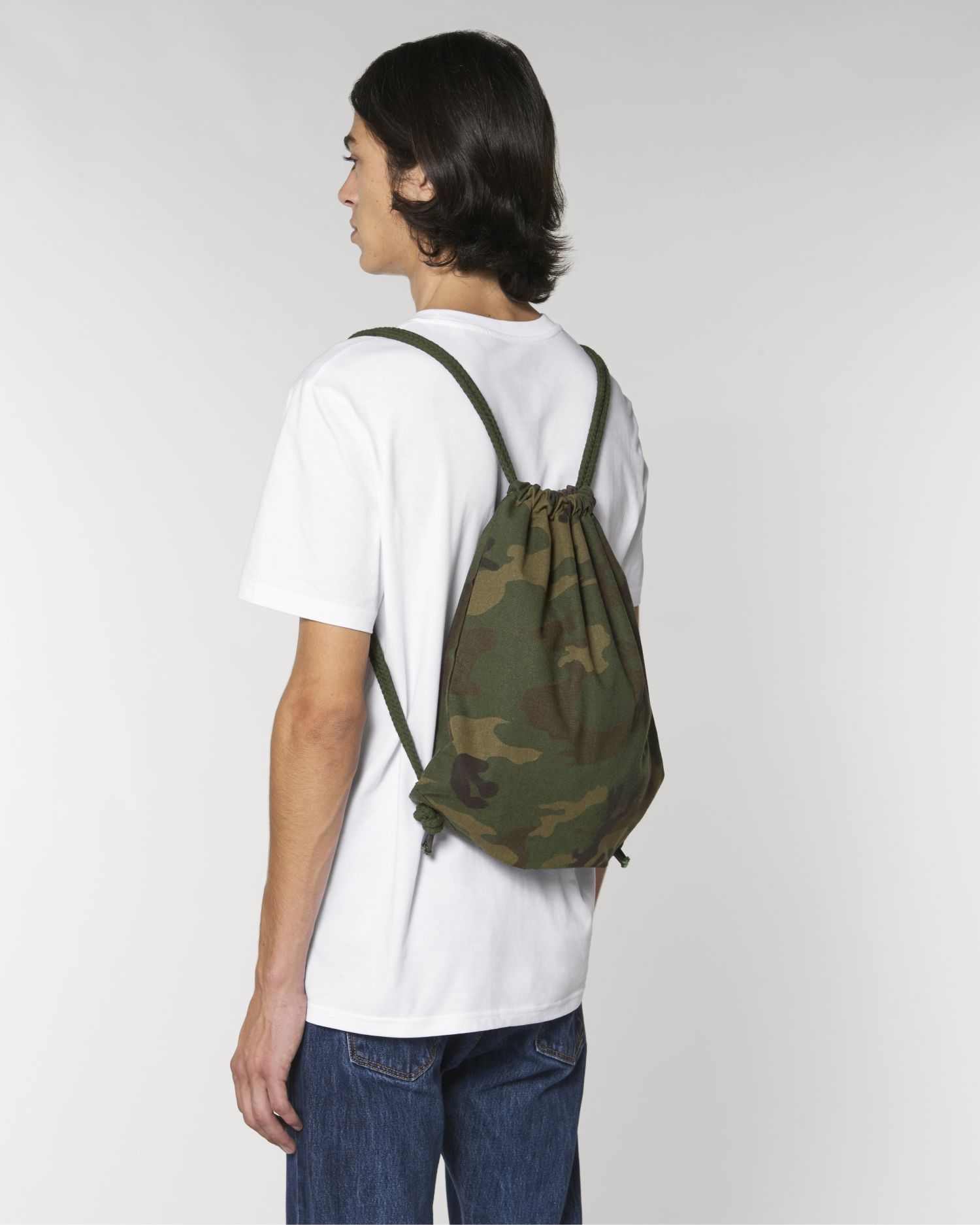 Joven de espaldas con mochila de saco camuflada verde y camiseta blanca sobre fondo neutro