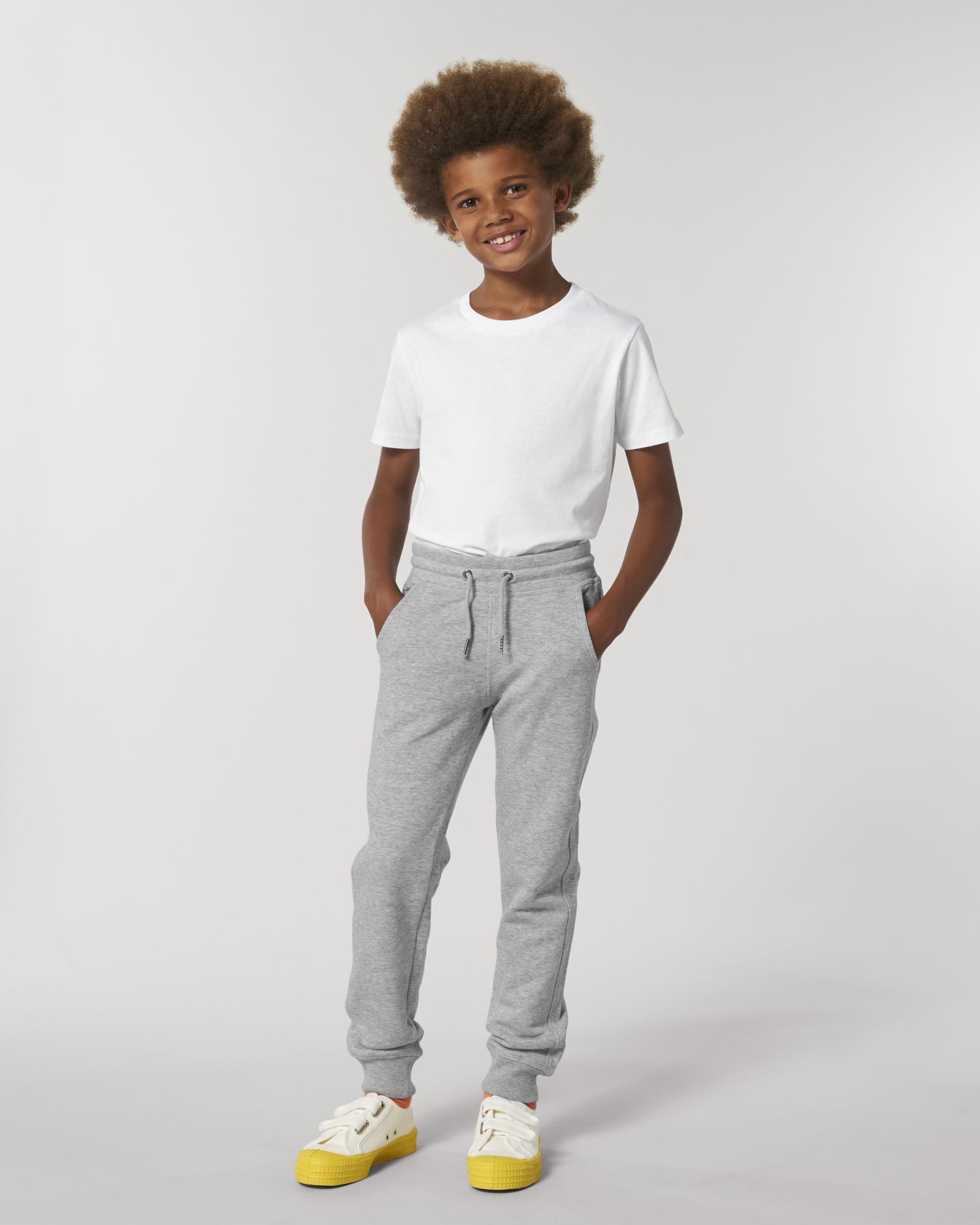 Niño sonriente con camiseta blanca, pantalones de chándal grises y zapatillas con suela amarilla sobre fondo neutro.