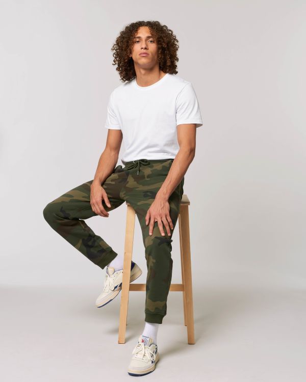 Hombre joven de cabello rizado sentado en un taburete con camiseta blanca y pantalones de camuflaje verde.