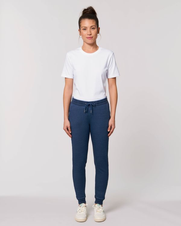 Mujer de pie con camiseta blanca, pantalones deportivos azules y zapatillas blancas sobre fondo gris claro