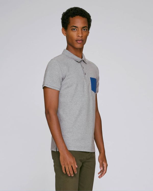 Hombre joven de pie con polo gris de bolsillo azul y pantalones verdes sobre fondo neutro.