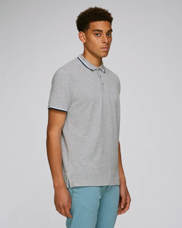 hombre joven de pie con polo gris de ribete blanco y azul y pantalones azules claros sobre fondo neutro