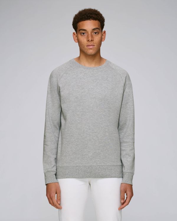 Hombre joven con sudadera gris lisa y pantalones blancos sobre fondo neutro.