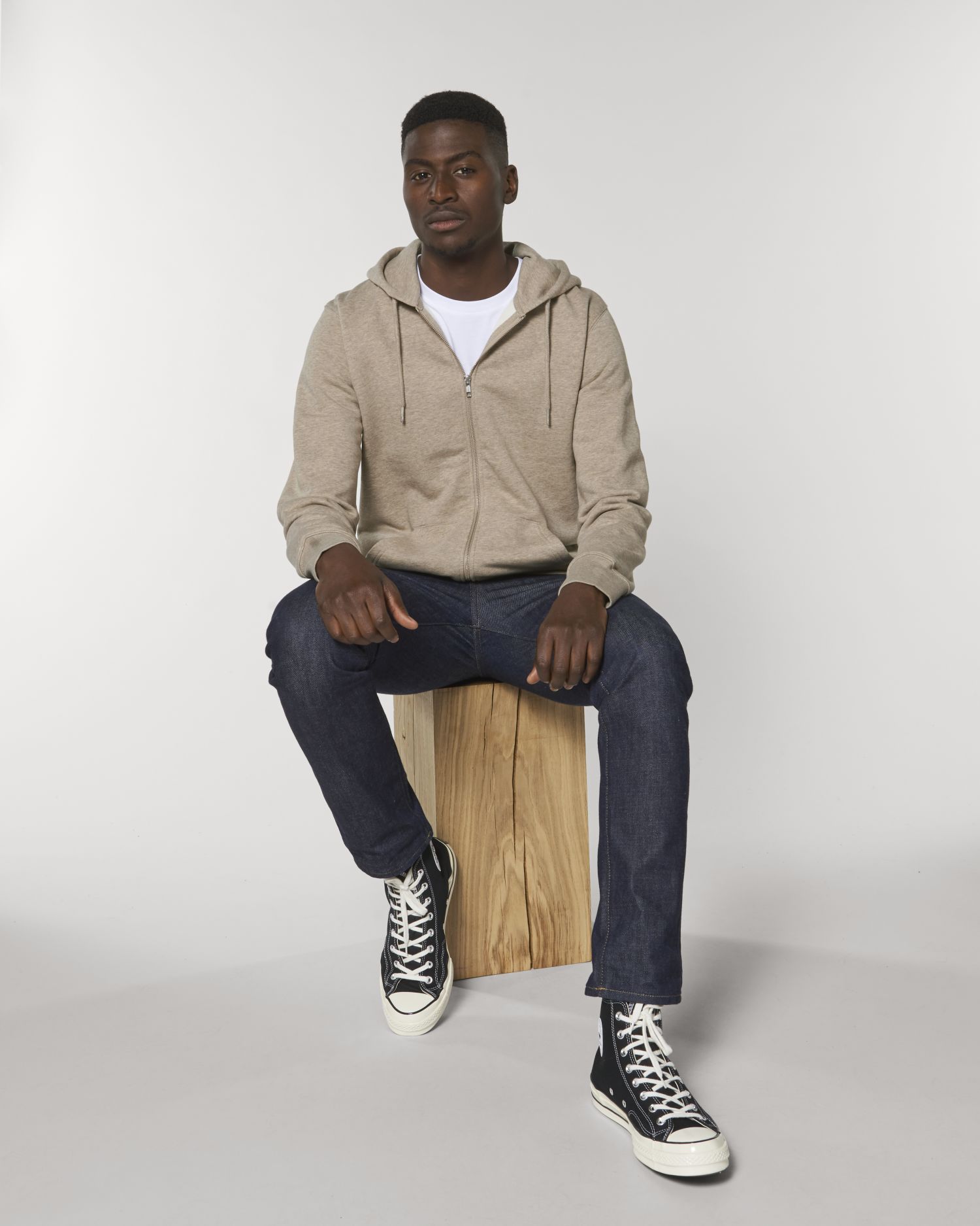 Hombre joven con sudadera beige y zapatillas negras sentado en un bloque de madera sobre fondo blanco