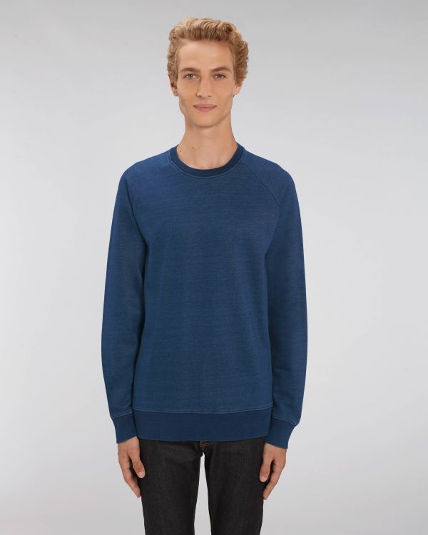 hombre de pie con sudadera azul y vaqueros oscuros sobre fondo neutro