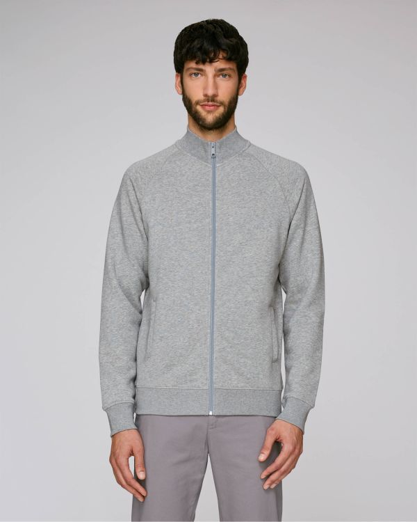 Hombre de pie con chaqueta deportiva gris de cremallera y pantalones claros sobre fondo neutro