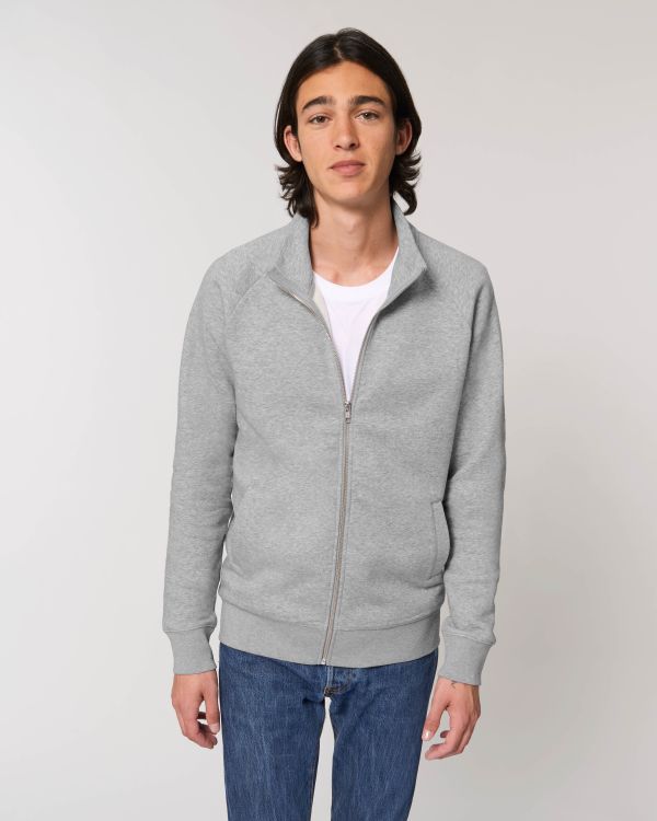 Joven con chaqueta gris con cremallera, camiseta blanca y vaqueros sobre fondo neutro.