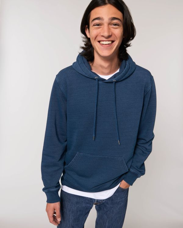Joven sonriente con sudadera azul de capucha y vaqueros sobre fondo blanco.