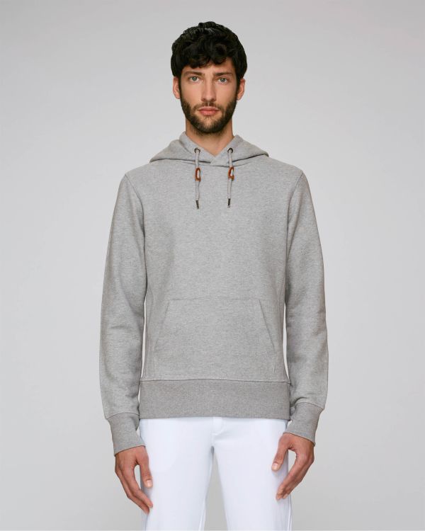 Hombre joven de pie con sudadera gris con capucha y bolsillo frontal sobre fondo claro.