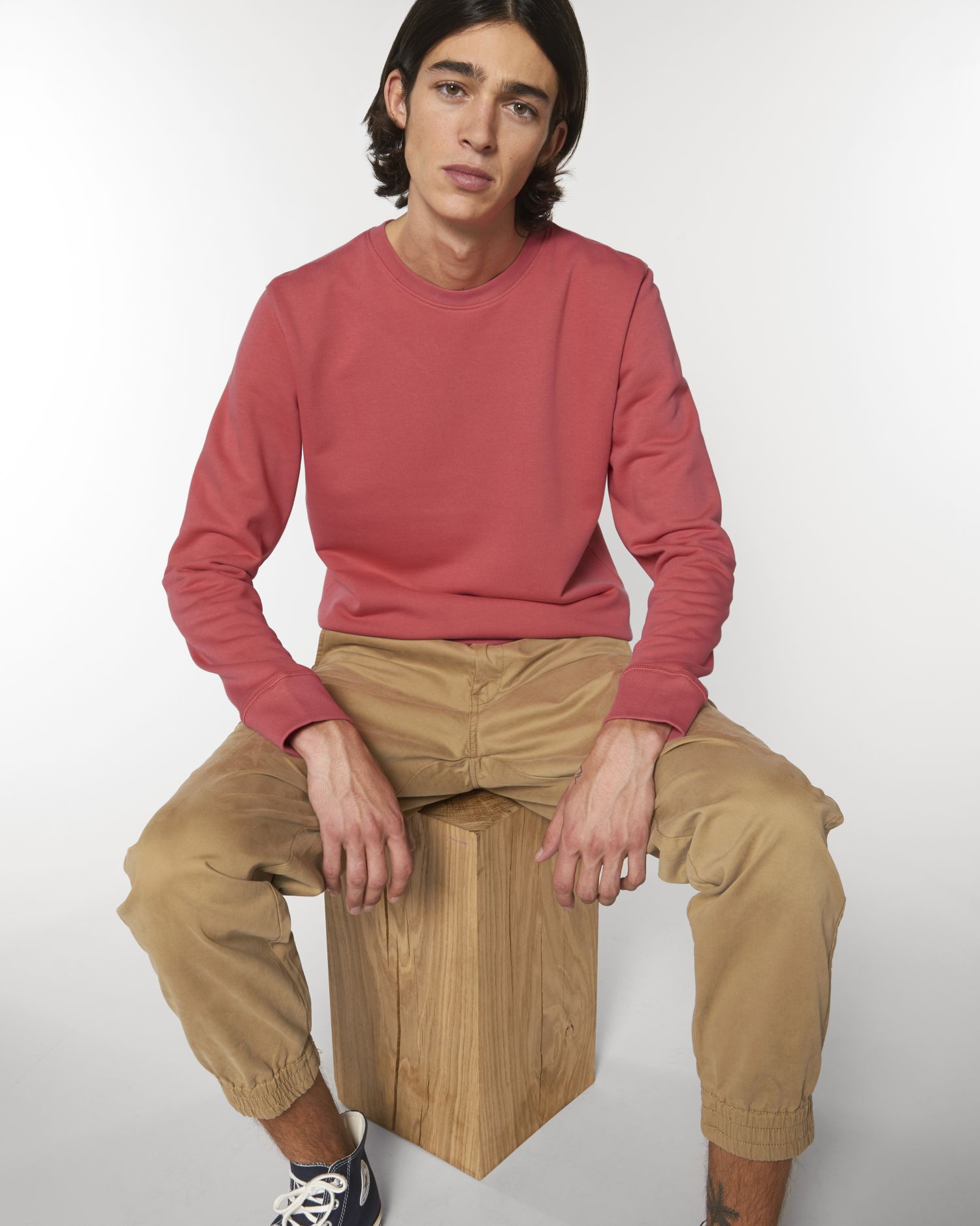 Hombre joven sentado en un bloque de madera con sudadera rosa coral y pantalones beige sobre fondo blanco