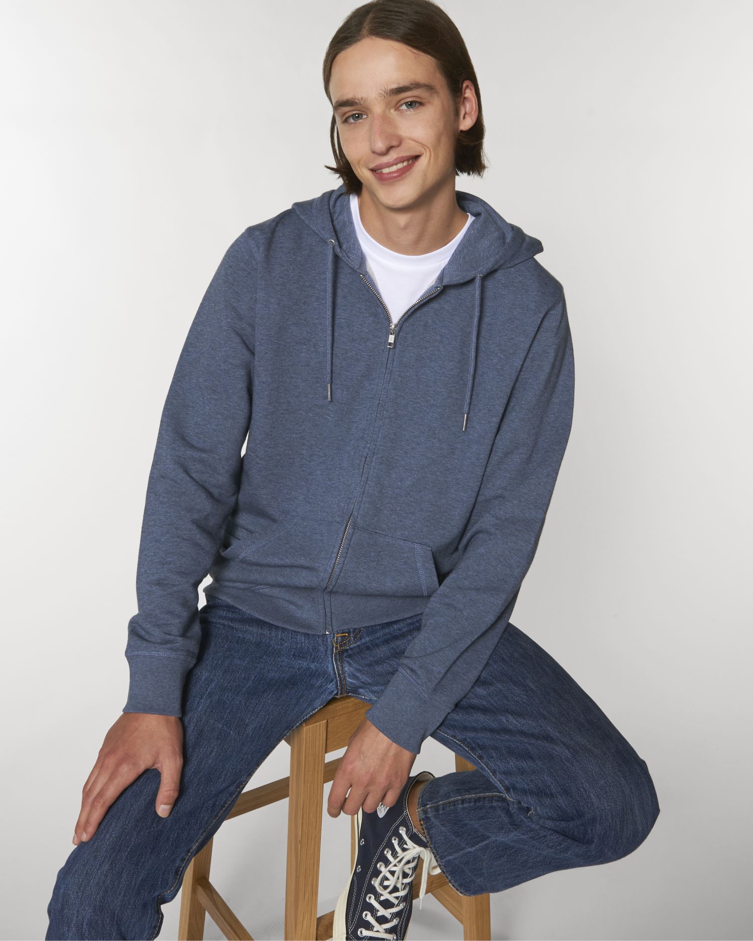 Joven sentado en un taburete con sudadera azul de cremallera, vaqueros y zapatillas deportivas sobre fondo blanco