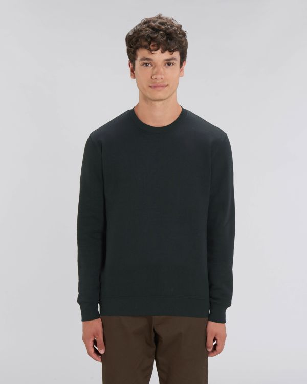 Joven con jersey negro de manga larga y pantalones marrones sobre fondo neutro.