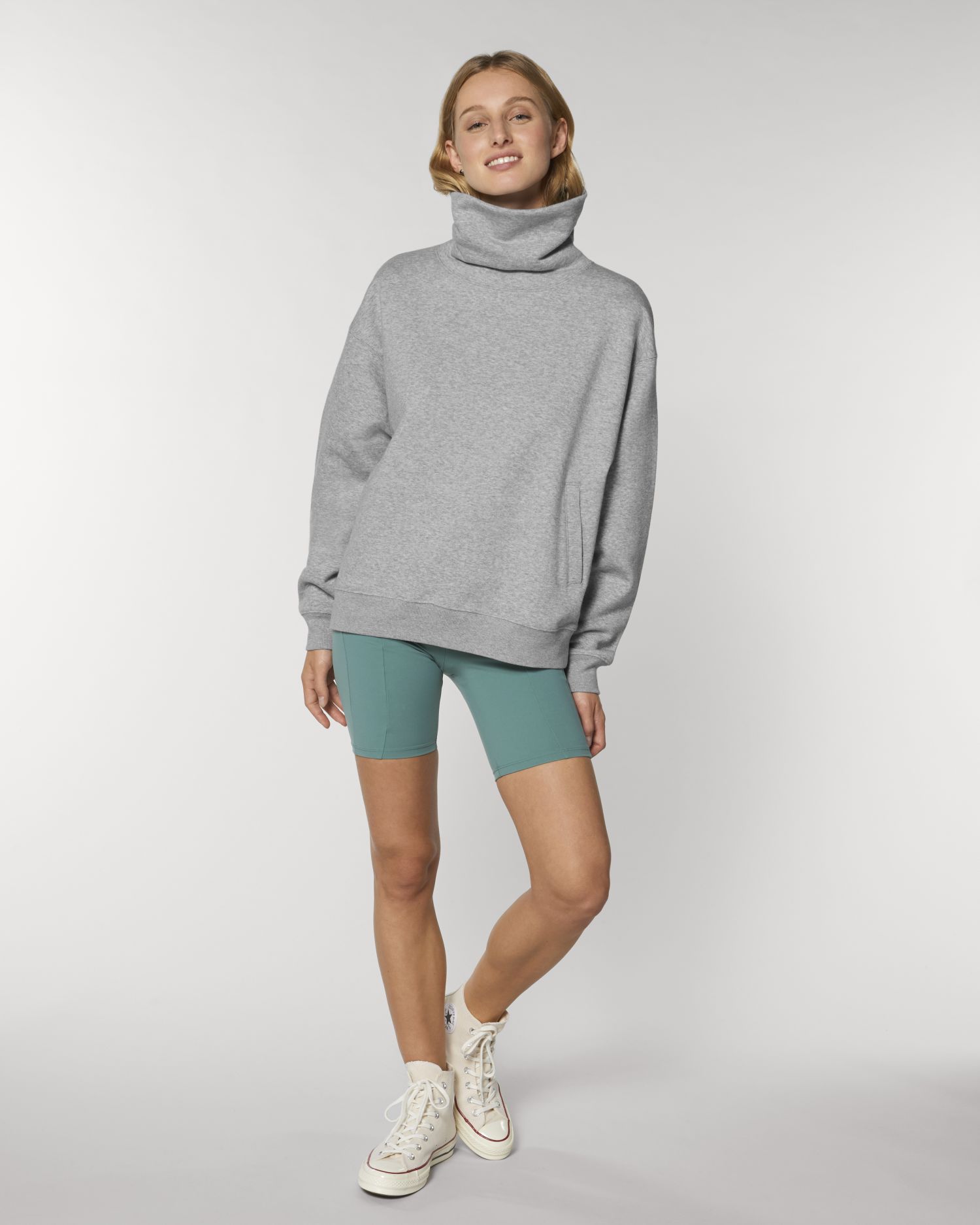 Mujer posando con sudadera gris de cuello alto, mallas cortas verdes y zapatillas blancas sobre fondo neutro