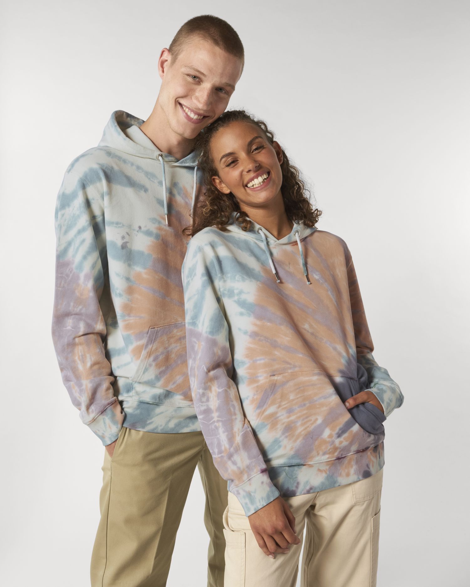 Pareja joven sonriendo con sudaderas con capucha de estampado tie-dye sobre fondo neutro.