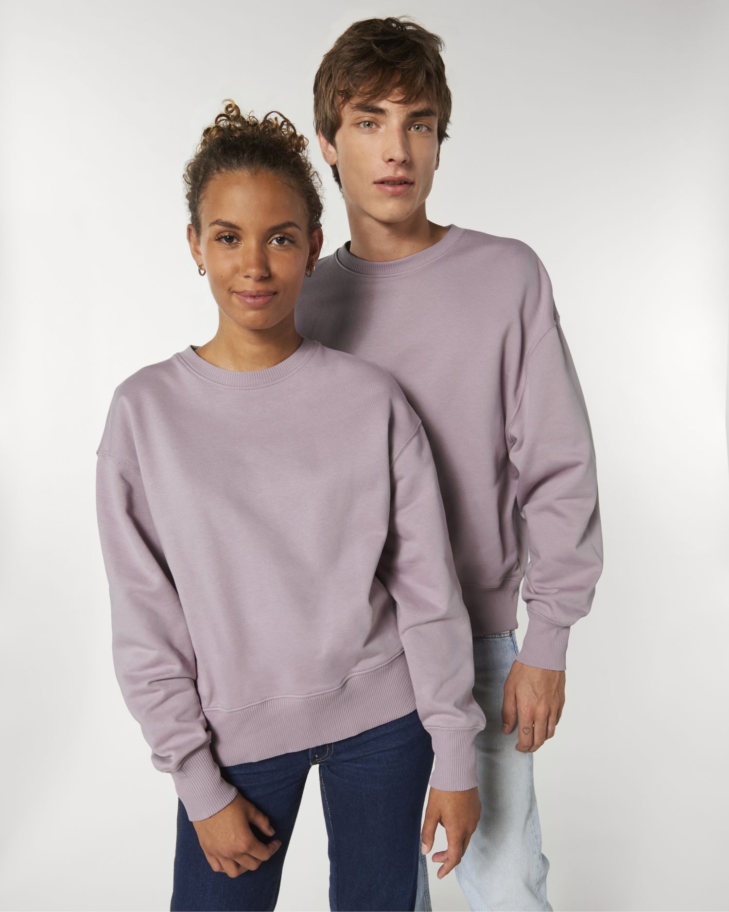 Dos jóvenes de pie con sudaderas malva lisas sobre fondo neutro.