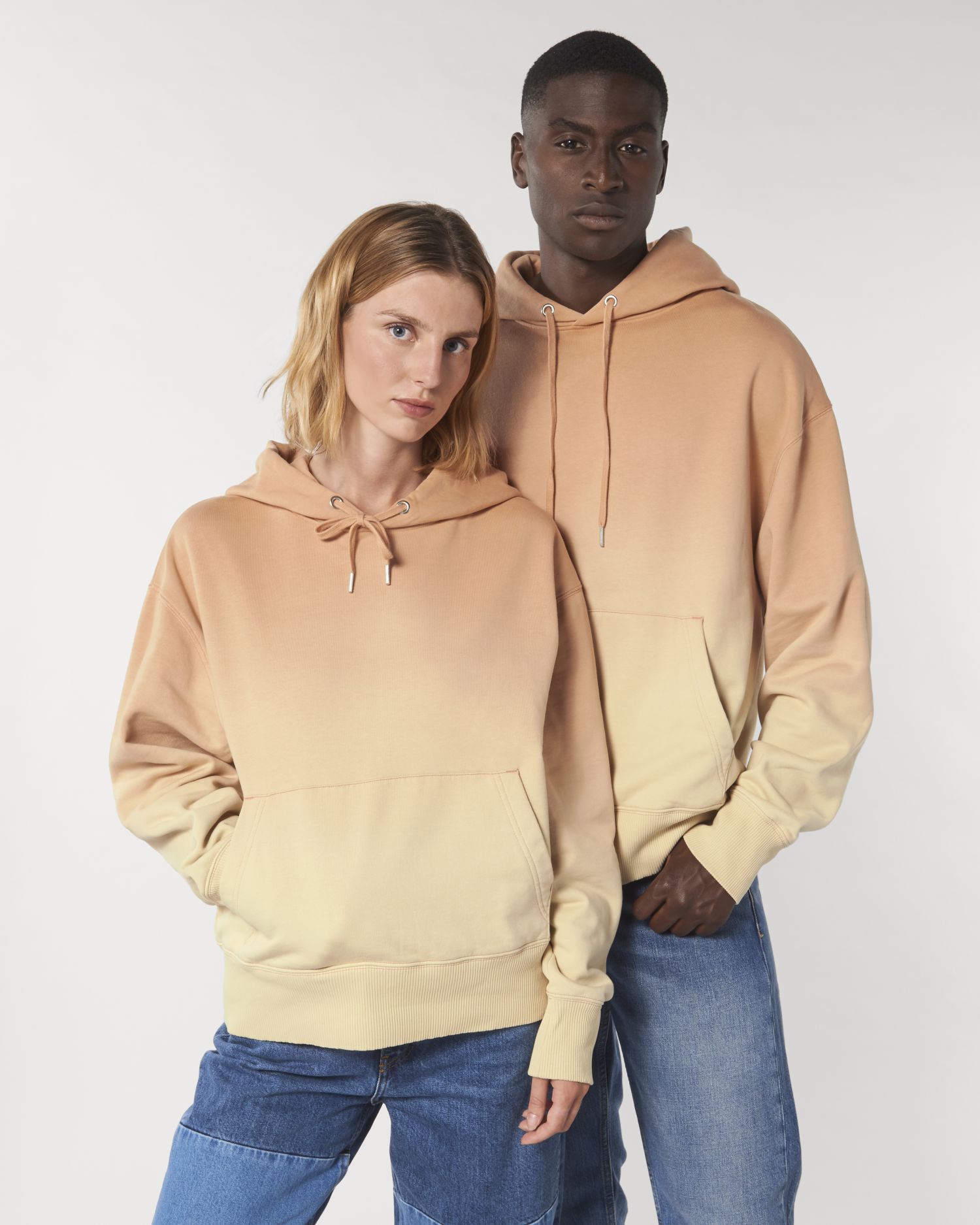Pareja de modelos con sudaderas con capucha degradadas en tonos melocotón y vaqueros sobre fondo blanco.