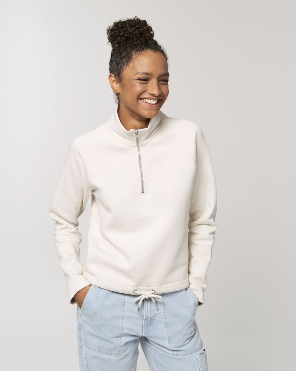 Mujer joven sonriente con sudadera blanca de media cremallera y vaqueros claros, de pie con las manos en los bolsillos sobre fondo neutro.