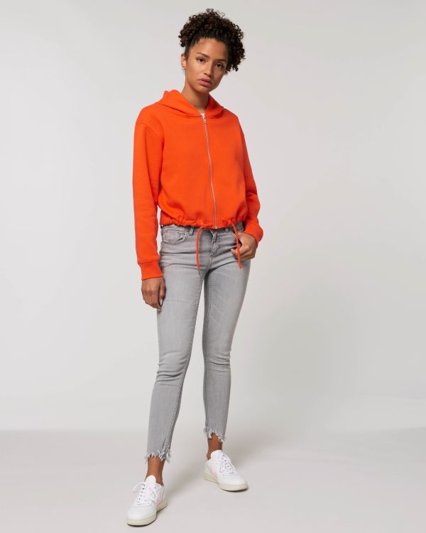 Mujer joven con sudadera naranja con capucha y cremallera, vaqueros grises ajustados con bajos deshilachados y zapatillas blancas sobre fondo neutro