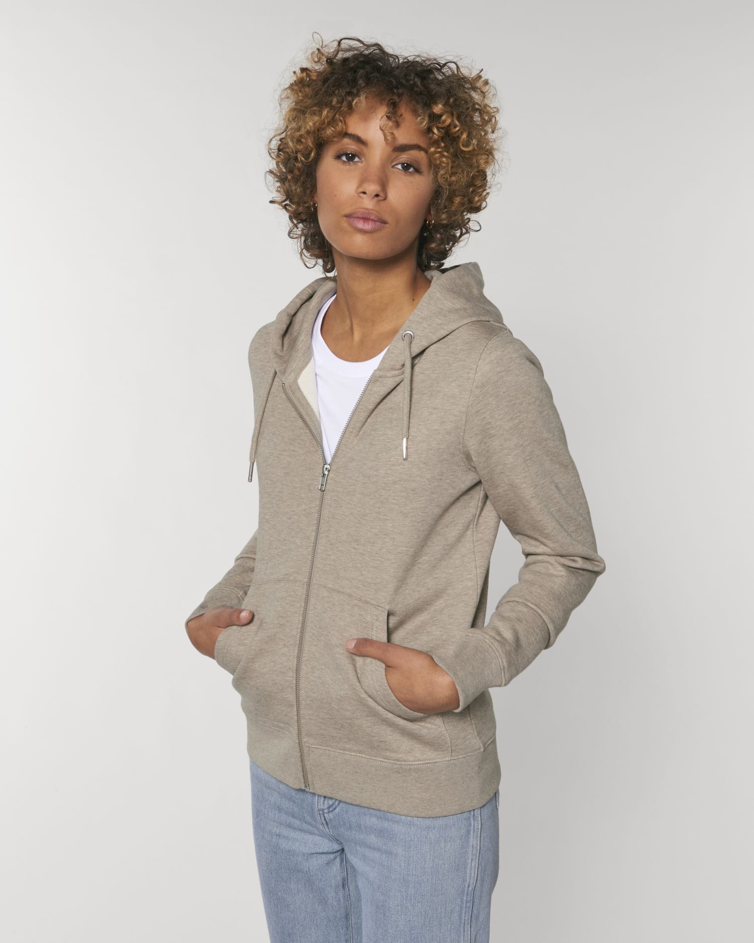 Mujer con sudadera beige con capucha y vaqueros claros posando en estudio con las manos en los bolsillos