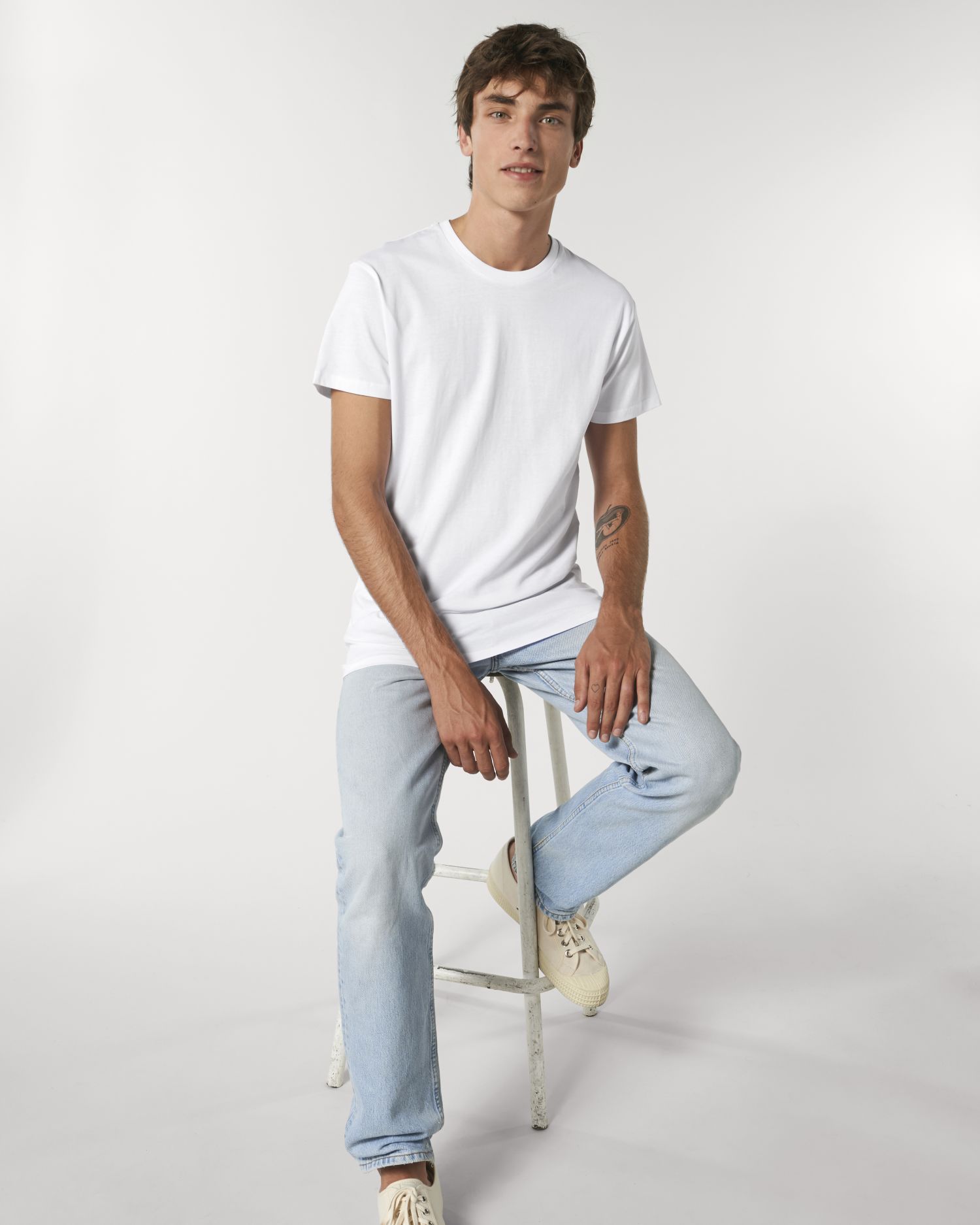 Joven sentado en un taburete con camiseta blanca, vaqueros claros y zapatillas sobre fondo neutro