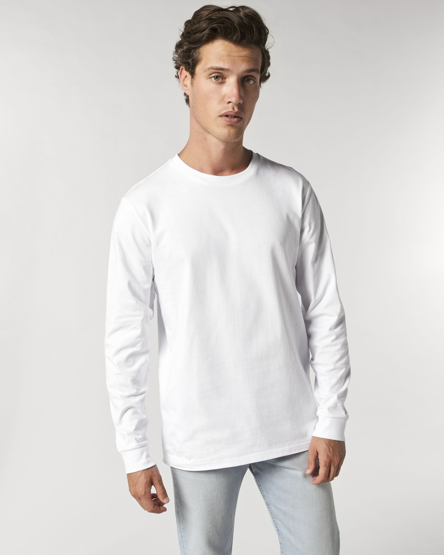 Hombre joven con camiseta blanca de manga larga y vaqueros claros sobre fondo neutro