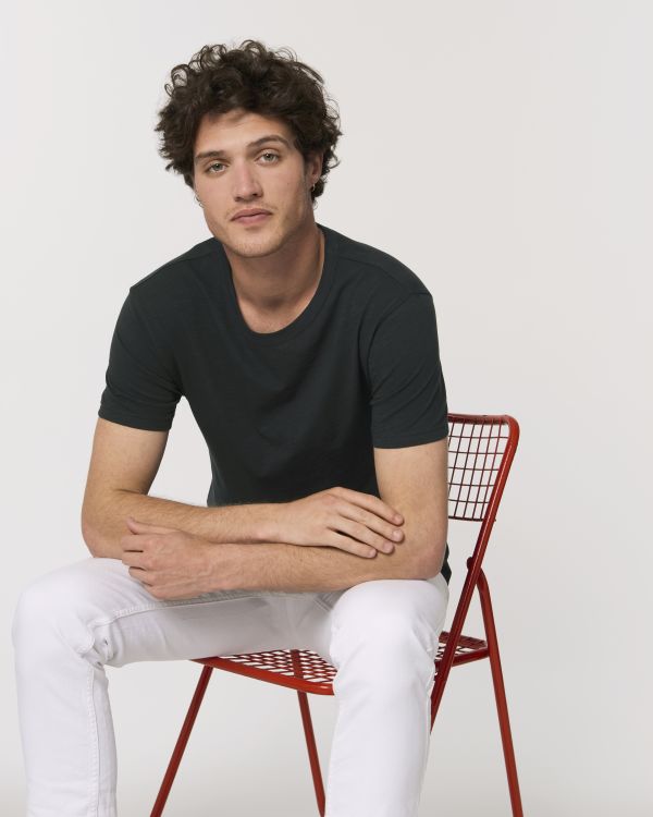 Hombre joven sentado en silla metálica roja con camiseta negra y pantalón blanco sobre fondo claro