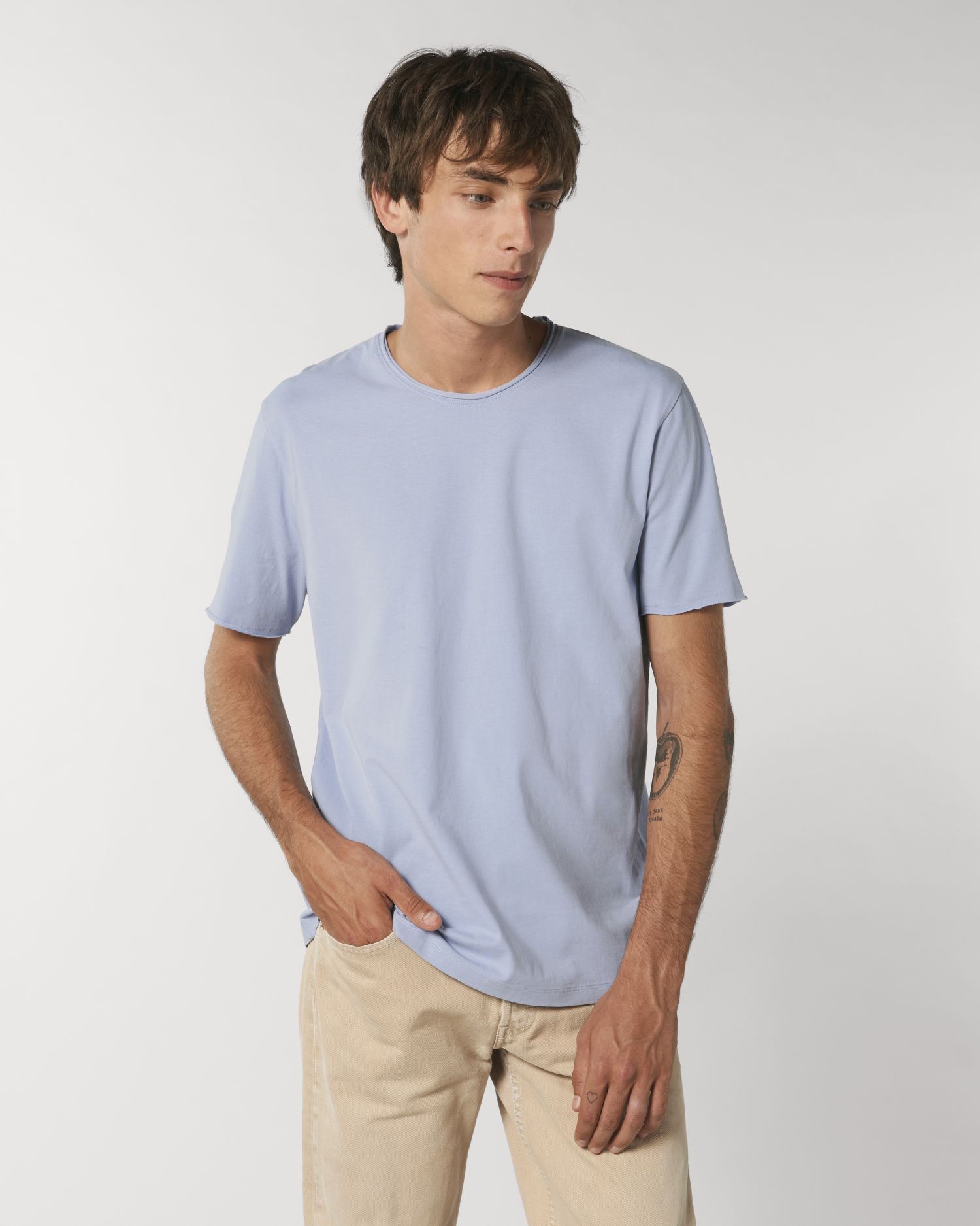 Hombre joven de pie con camiseta azul claro y pantalones beige sobre fondo blanco.