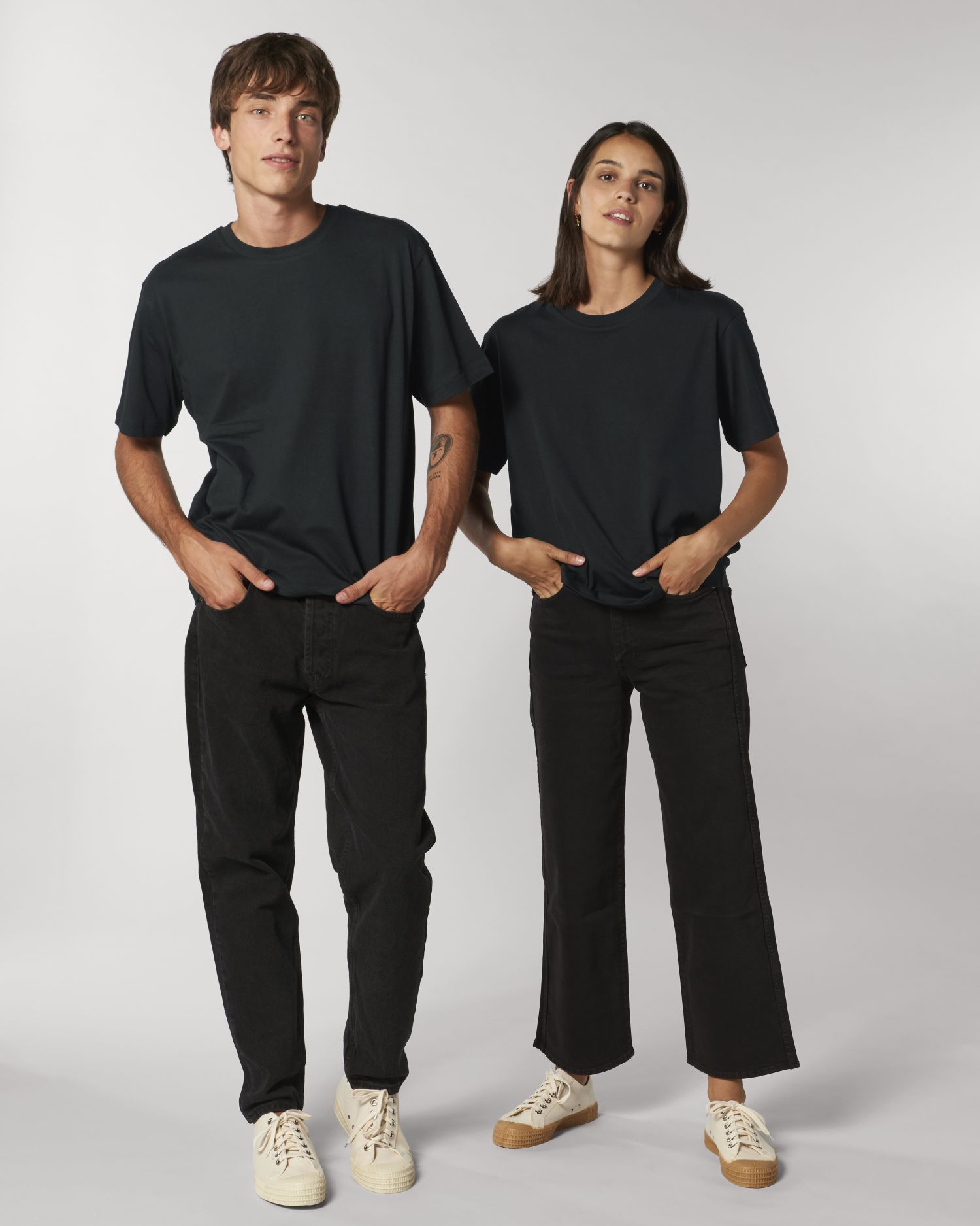Pareja posando con camiseta negra de manga corta, vaqueros negros y zapatillas blancas sobre fondo gris claro