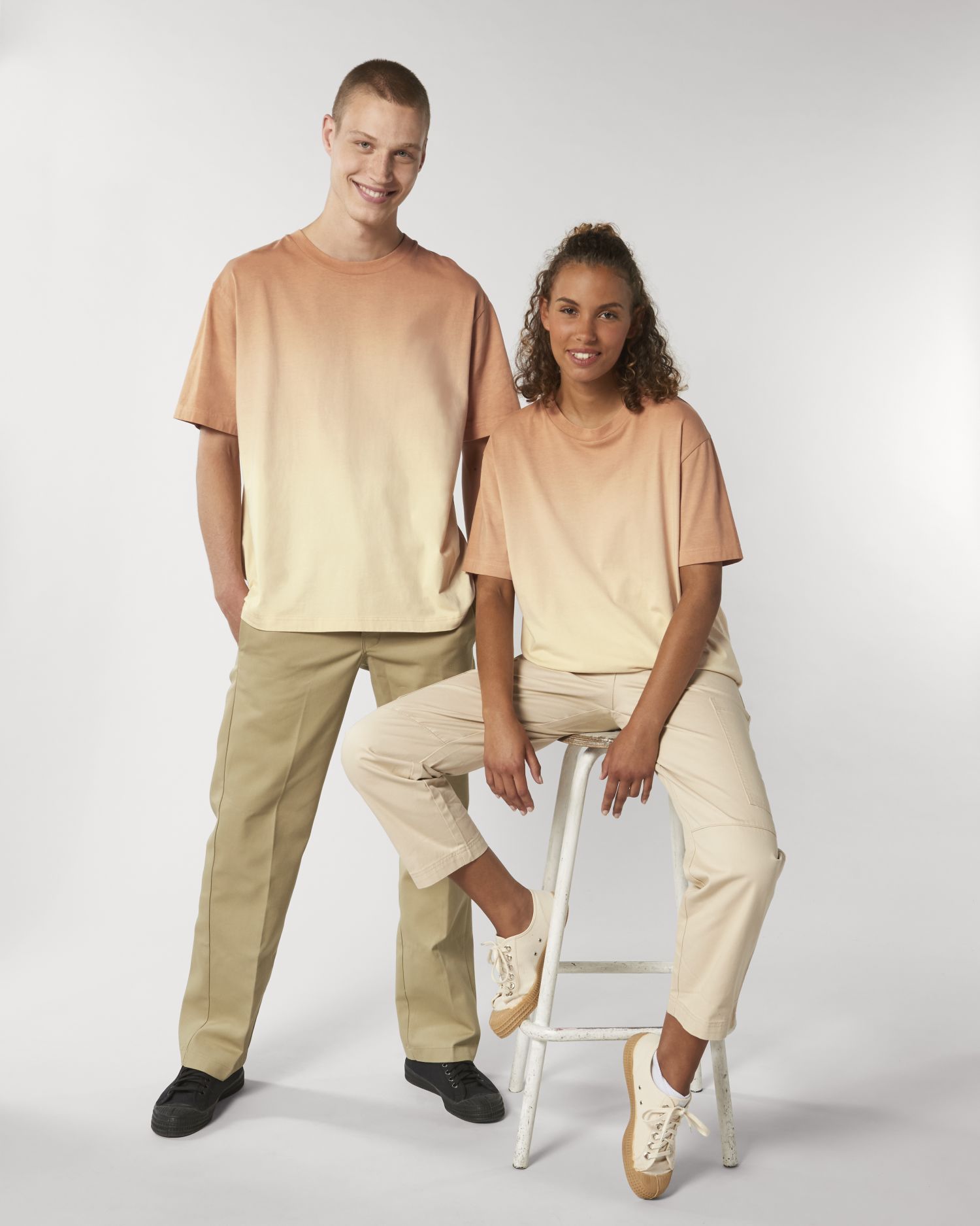 dos jóvenes, un chico de pie y una chica sentada en un taburete, vistiendo camisetas degradadas en tonos salmón y pantalones claros frente a fondo blanco