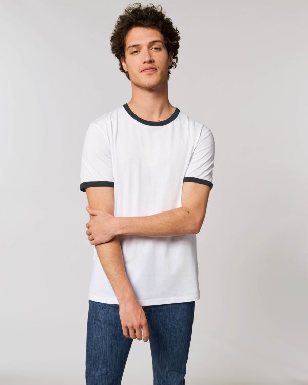 Hombre joven con camiseta blanca de cuello y mangas negras y vaqueros sobre fondo neutro.