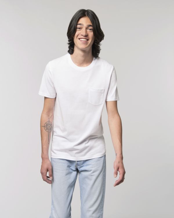 Joven sonriente de pie con camiseta blanca, vaqueros claros y tatuaje de rosa de los vientos en el antebrazo sobre fondo neutro.