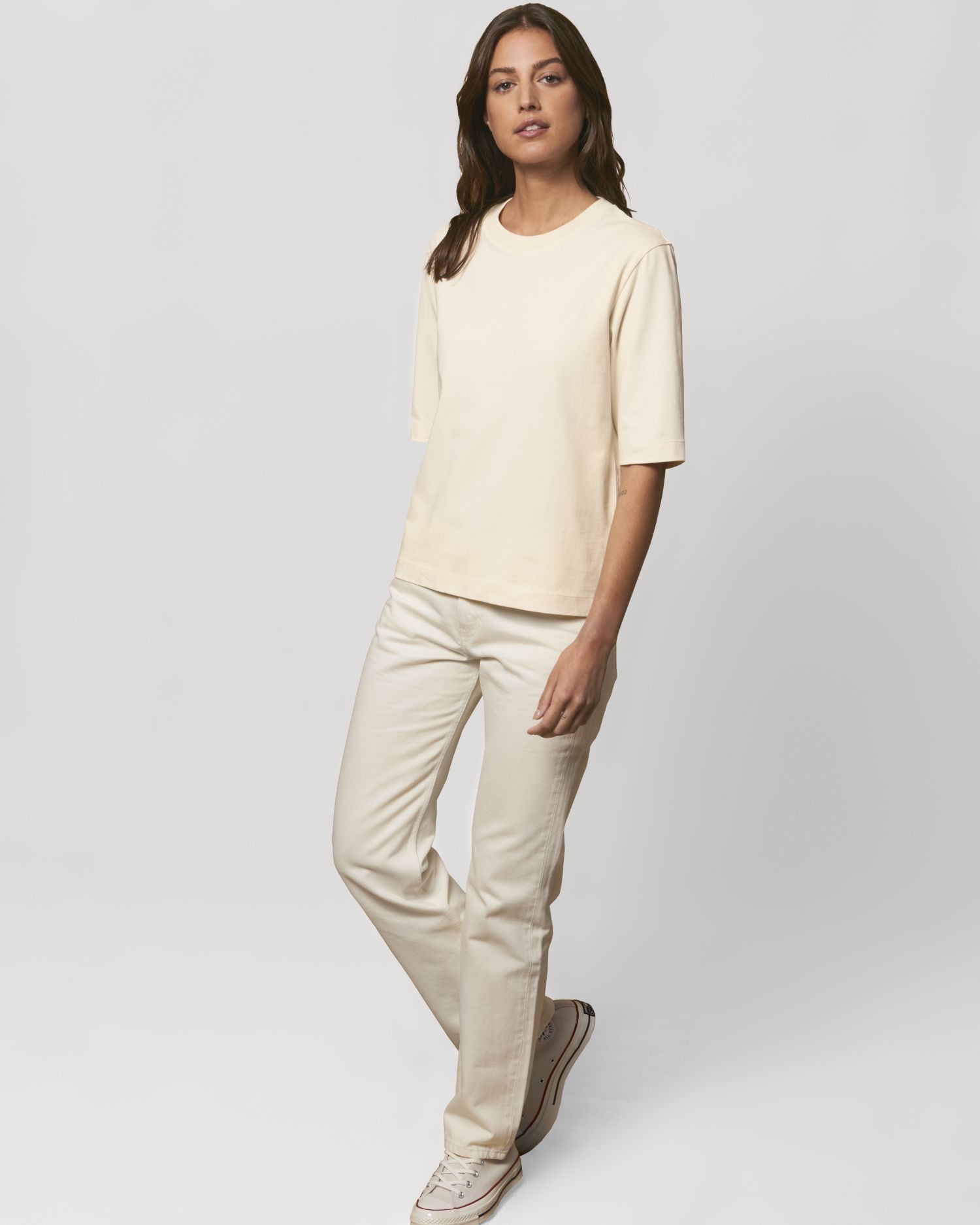 Mujer de pie con camiseta de algodón crema, vaqueros claros y zapatillas blancas sobre fondo neutro.