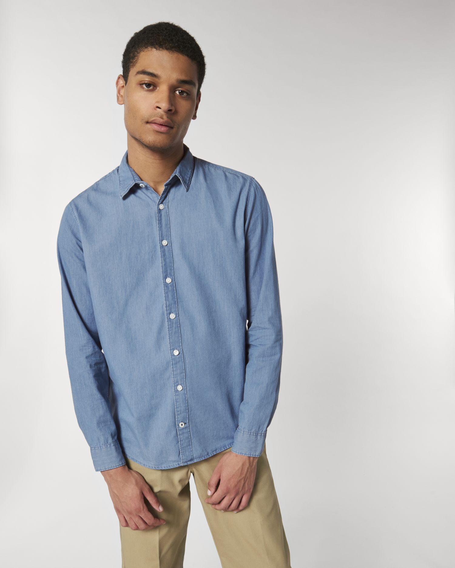 Hombre joven con camisa vaquera azul claro y pantalones chinos beige sobre fondo neutro.
