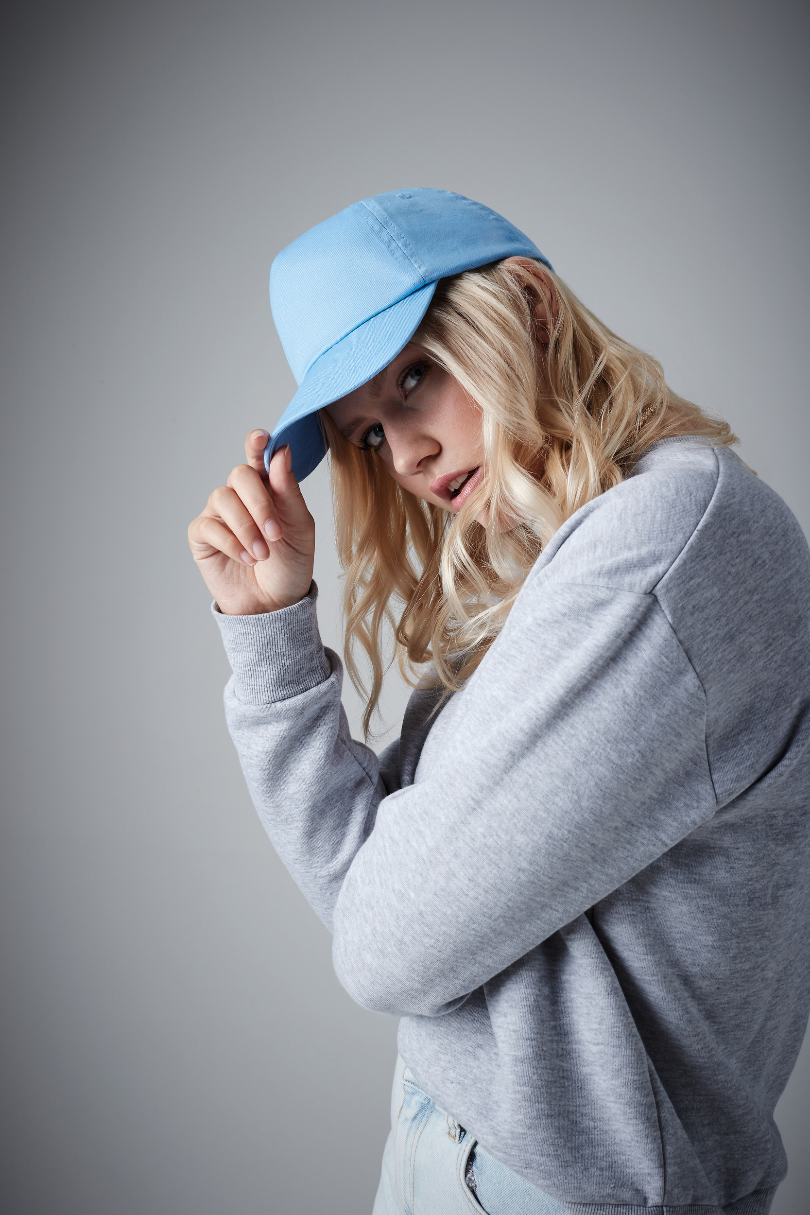 Mujer joven rubia con gorra azul y sudadera gris posando de perfil en estudio.