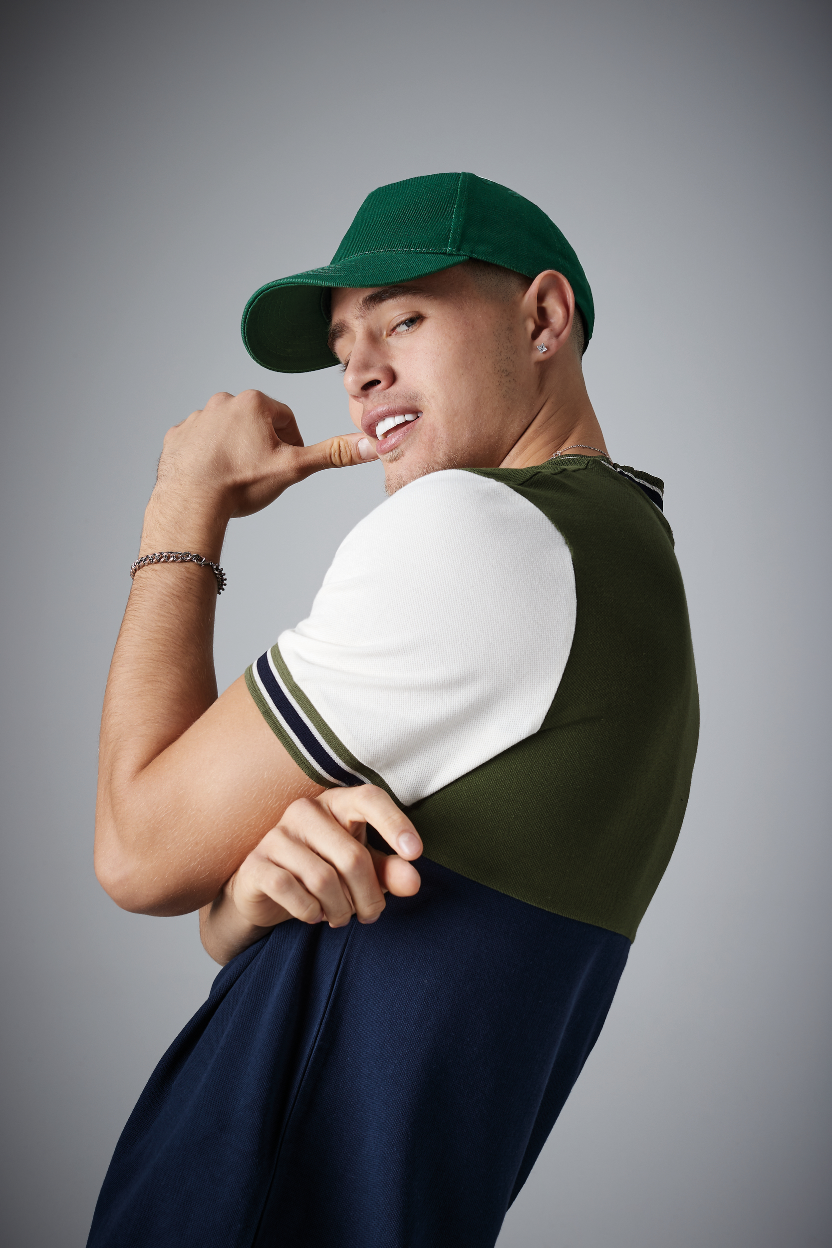 Joven con gorra verde y camiseta bicolor posando de perfil con actitud relajada.