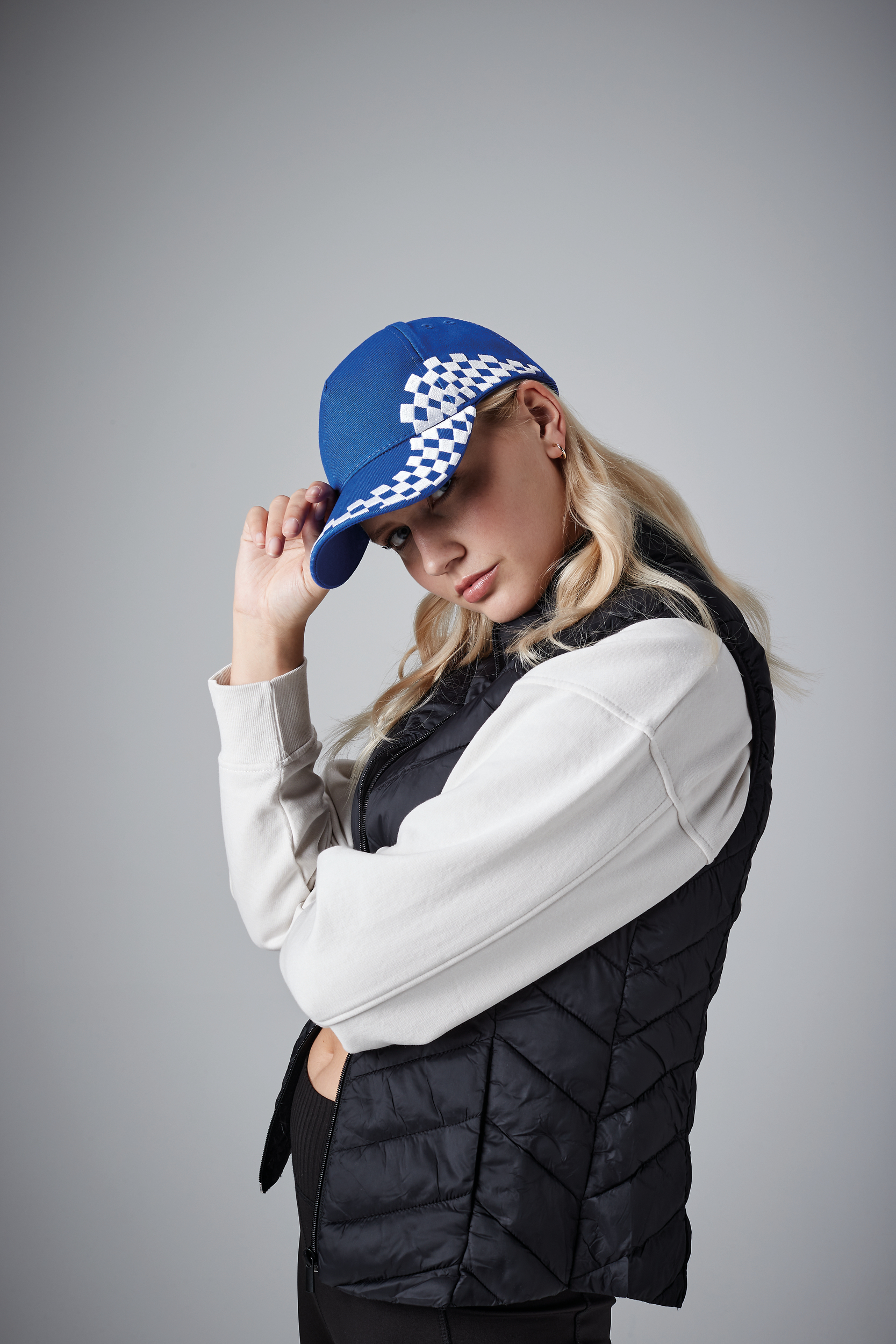 Mujer joven con gorra azul de cuadros blancos, chaleco negro acolchado y sudadera blanca sobre fondo gris.