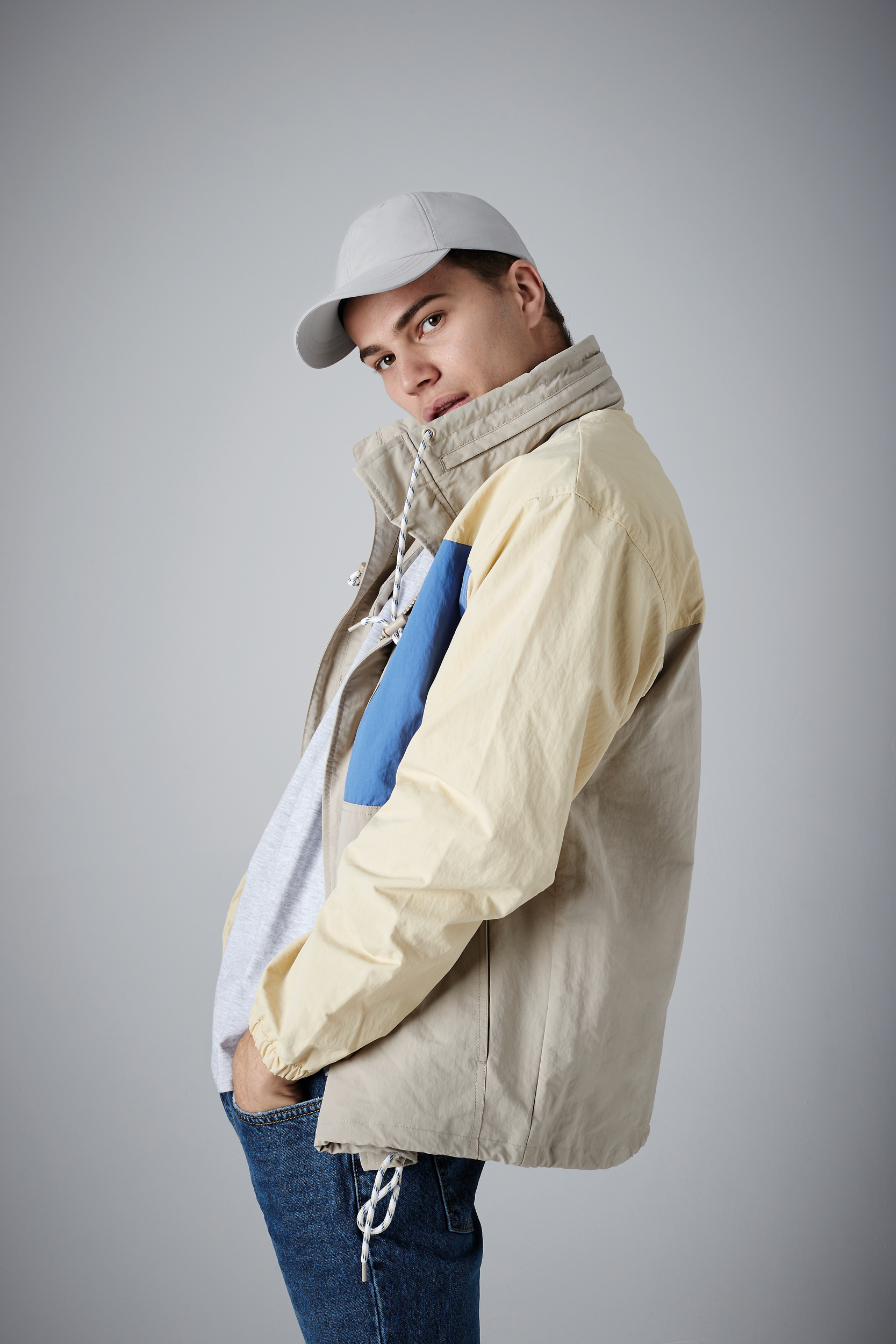 hombre joven con gorra y cazadora ligera de tonos beige y azul sobre fondo gris claro