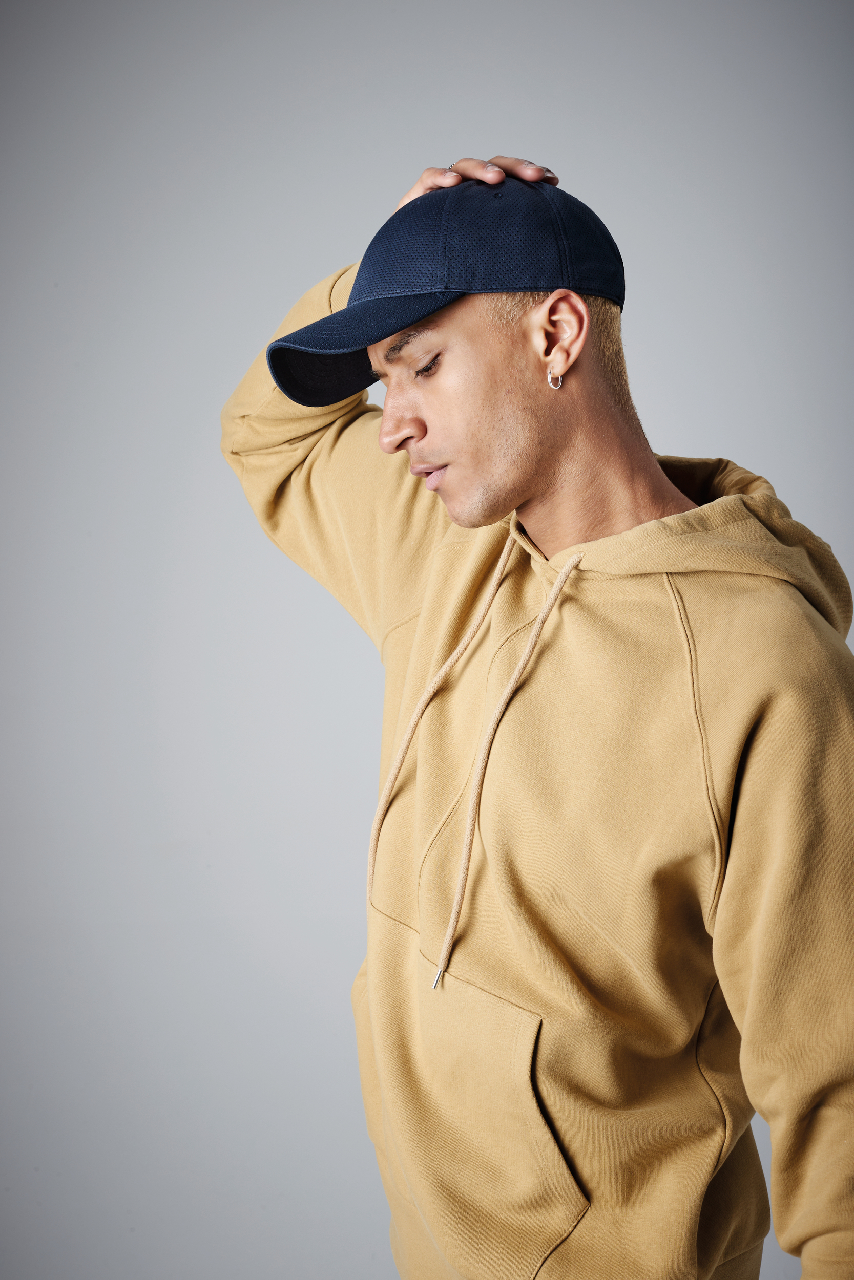 Hombre joven con sudadera beige y gorra azul marino, con la mano en la cabeza sobre fondo gris claro