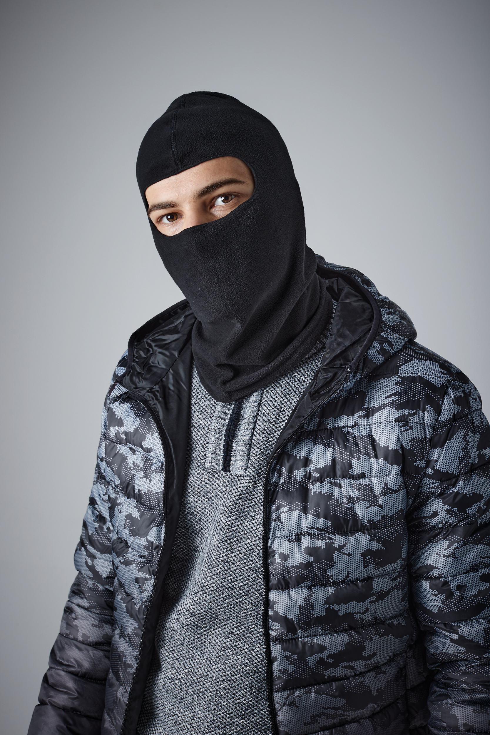 hombre con pasamontañas negro y chaqueta acolchada de camuflaje sobre fondo neutro