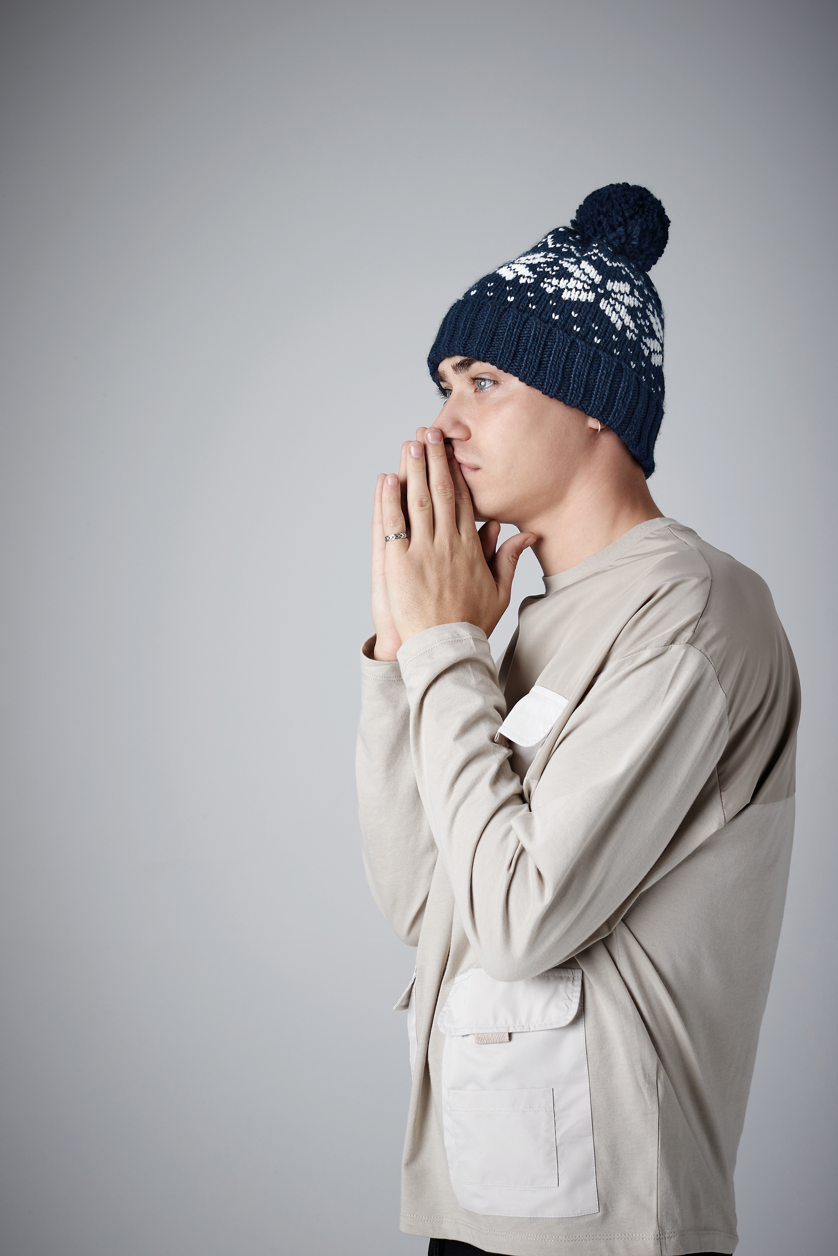 Hombre joven de perfil con gorro de punto invernal y manos juntas junto al rostro sobre fondo gris claro.
