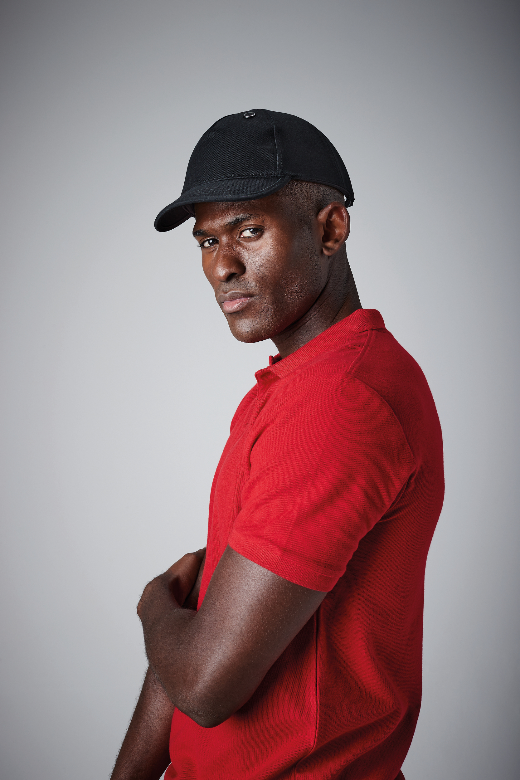 Hombre de piel oscura con gorra negra y camiseta roja de perfil sobre fondo gris mirada a cámara